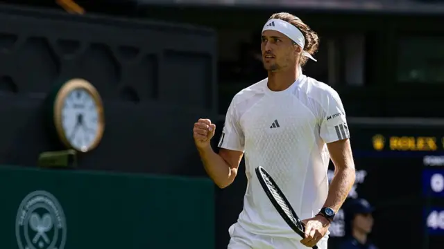 Zverev, wearing a white t-shirt and a white bandana with his hair tied in a bun, holds his racquet in his left hand and clenches his right fist