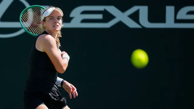 McNally, wearing a black dress and white visor, follows through after a backhand with her green racquet behind the shoulder and the yellow ball blurred in the forehand on the right