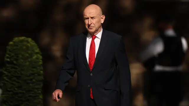 John Healey, a man, wearing a dark suit and red tie, walking into 10 Downing Street.