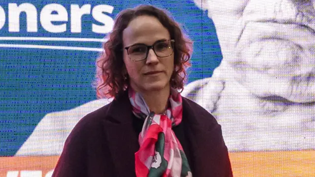 A woman with shoulder-length brown, curly hair. She is wearing glasses, a claret overcoat and a pink scarf.