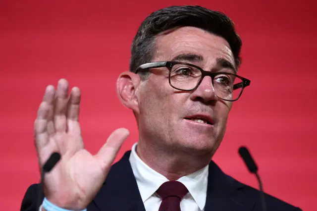 Andy Burnham, a white man with dark hair and glasses, in a dark suit and maroon tie. He is speaking into a microphone and his hand is raised.