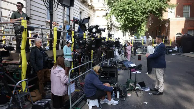 Crowds of people with cameras and microphone stand on Downing Street, metal barriers are in place.