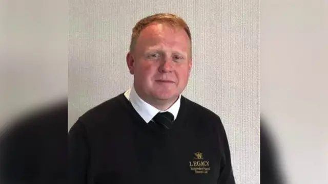 Robert Bush has short ginger hair. He is wearing a white shirt, a black tie and a black jumper.