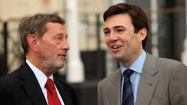 David Blunkett (L) in a dark suit, white shirt and red tie with blue polka dots speaks to a young Andy Burnham (R) in a grey suit, light blue shirt and striped dark blue tie
