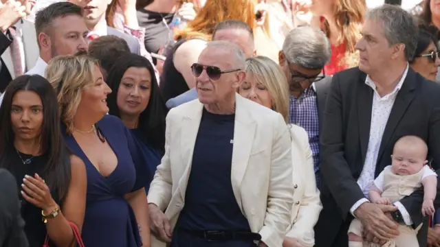 Reid in the middle of a crowd with sunglasses on and a white blazer. A man holds a baby on one side, three women flank his other side