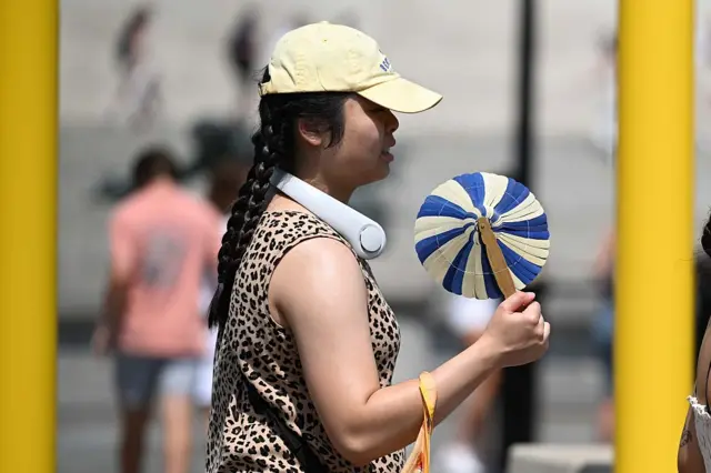 A woman walking with a hat on, a fan in her hand, and a portable fan round her neck