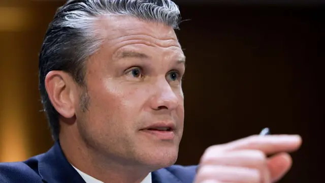 Closeup of Hegseth as he holds his hand up, pinching his forefinger and his thumb