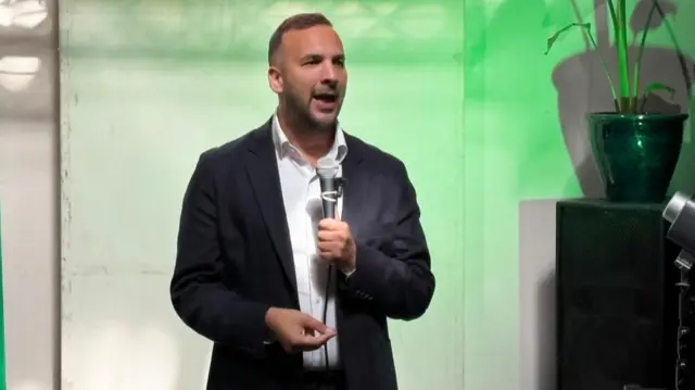 Green Party Leader Zack Polanski holding a microphone in front of a green lit background.