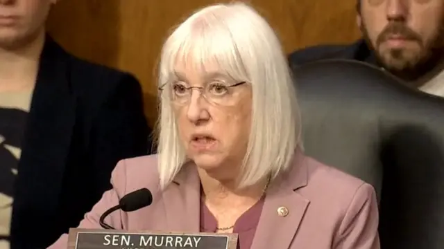 Murray speaks into a small mic as she sits on a black leather chair in front of two people sitting behind her