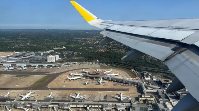 An aircraft wing with a yellow tip. On the ground below is a large airport terminal with several aircraft parked up.