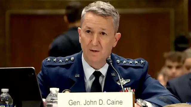 Caine speaks into a mic that's propped up on a desk in front of him as he sits