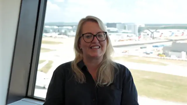 Jo Crawford smiling next to a big window. An airfield is visible behind her.