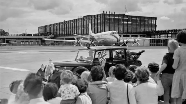 An old aeroplane beside a steel and glass terminal in a black and white image. A 1950s Rolls Royce with a young Elizabeth II and Prince Phillip in it drives past a large crowd of people on the airfield.