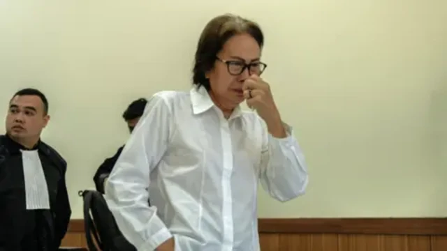 A woman wearing a white shirt and black-rimmed glasses looks emotional and holds a tissue up to her nose