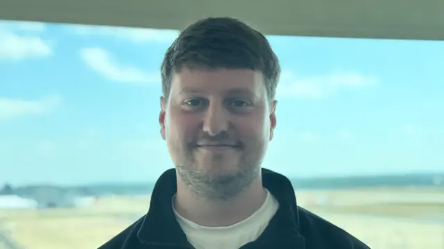 Oscar is smiling at the camera, standing in front of the airfield and he's wearing a black fleece and white t-shirt.