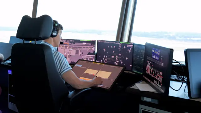 A woman in a headset sits in front of a large array of computer screens. She is in a room with large, bright windows.