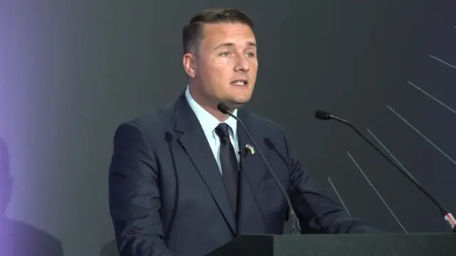 Wes Streeting standing in front of two microphones. He is wearing a black suit