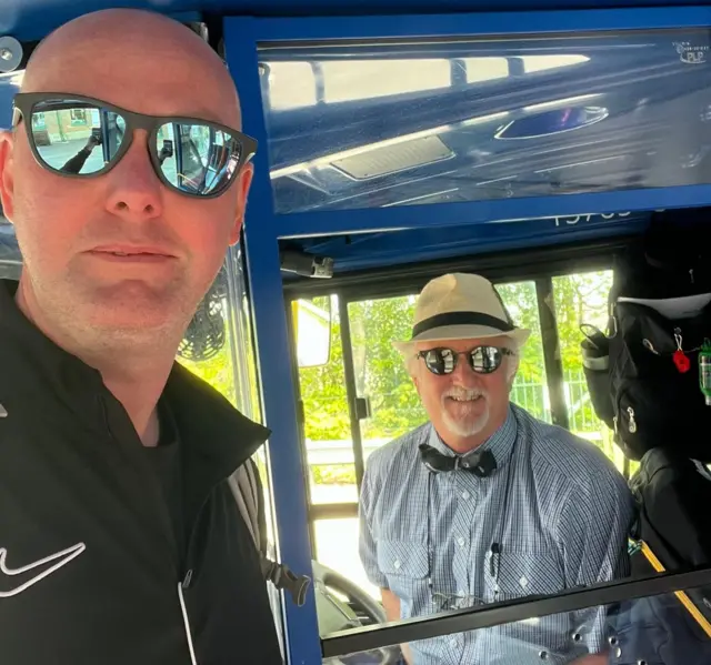 James taking a selfie on a bun with a bus driver, with both wearing sunglasses