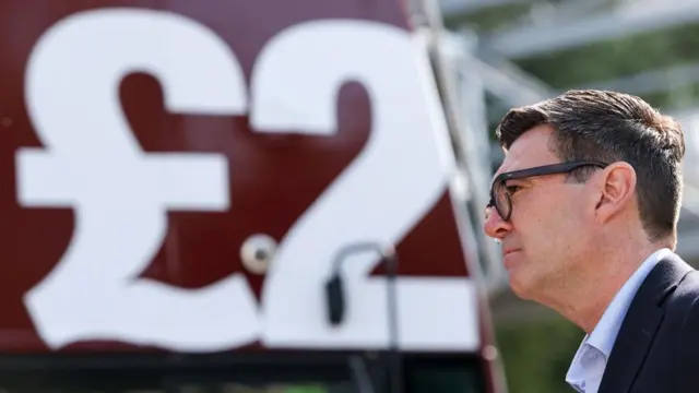 A side profile of Andy Burnham wearing a suit with a bus in the background. A wrap saying "£2" is visible on the bus.