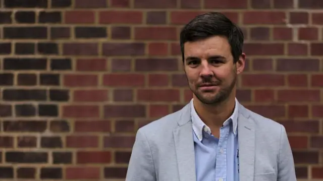 Seb Pelisse wears a white jacket and blue shirt, smiling
