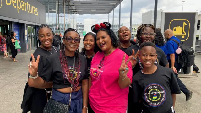 Leila Hall in a pink t-shirt, surrounded by multiple family members in matching black t-shirts. They are smiling and some are making the peace sign with their hands outside Gatwick Airport departures.