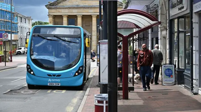 A blue Arriva bus arrives at a bus stop by shops in Royal Turnbridge Wells