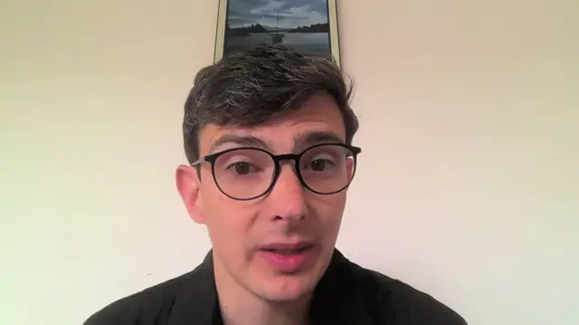 Alex Chapman sits instead of a white wall with a painting at the top. He is wearing a dark shirt and glasses.