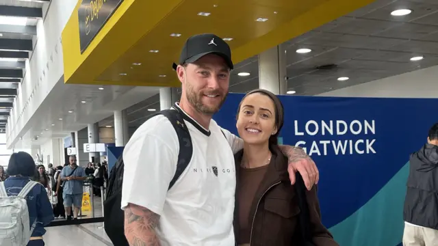 Louise and Dan with their arms around each other. They are in an airport terminal and a sign behind them reads London Gatwick