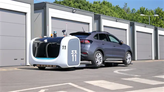 An SUV on a low flatbed, being pulled by some sort of robotic vehicle past some garages.
