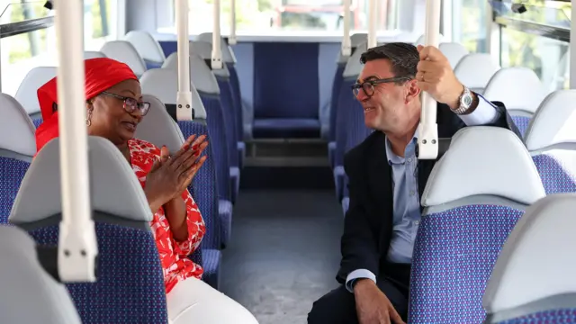 Andy Burnham sits on a bit wearing a suit next to Bemi Orojuogun who is wearing a bright red dress. The seats on the bus are blue.