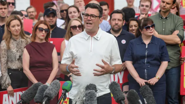 Andy Burnham speaks into media microphones with a team of people behind him