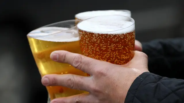 A man holds three glasses of beer in his hands, wearing a black quilted jacket.