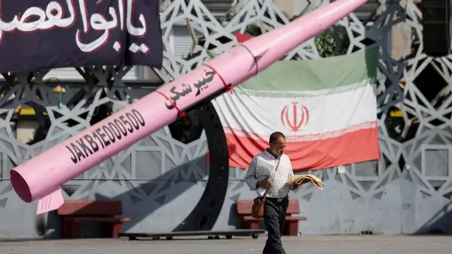 A man walks next to a symbolic mockup of an Iranian missile and an Iranian flag at Imam Hussein Square in Tehran, reading a newspaper and looking down