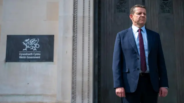 Rhun ap Iorwerth stands in a navy suit beside a sign that says "Welsh Government" in English and Welsh.