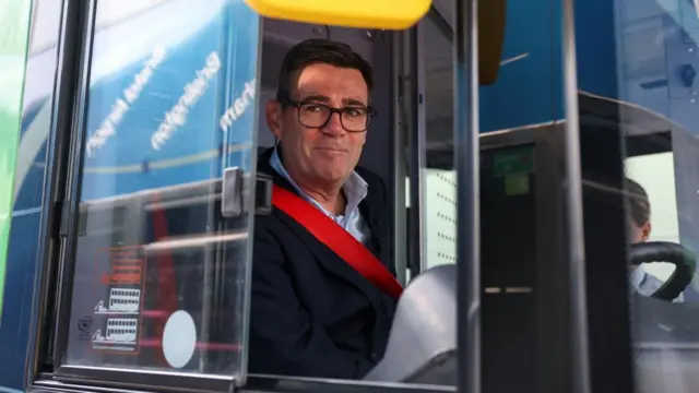 Andy Burnham sitting in the driver's seat on the bus putting on his seatbelt and seen through the window