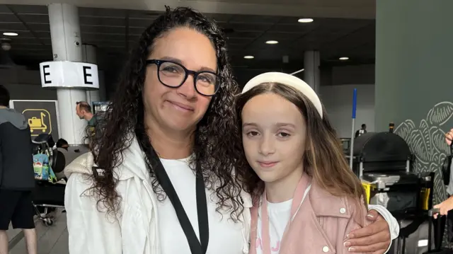 Medina stood with her arm around Kaci and smiling. People are pushing baggage trolleys through an airport terminal behind them.