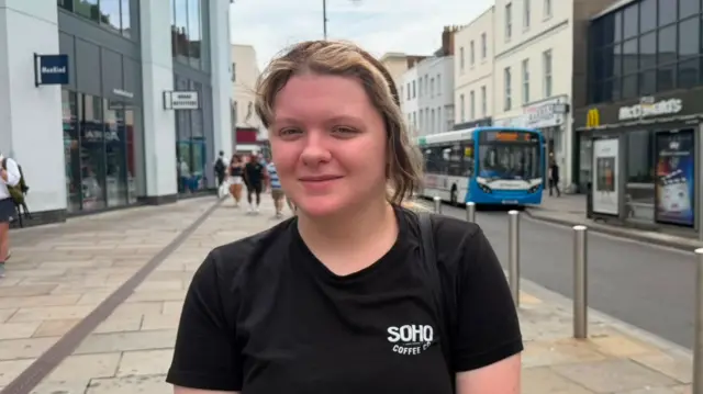 Woman wearing a black t-shirt reading 'Soho Coffee Co' stands and looks into the camera while standing on pavement next to a road. A bus is parked up in a bus stop in the background.