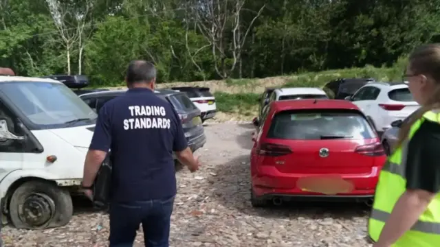 A woman in a high-vis jacket and a man in a dark shirt that reads 'trading standards' examine multiple cars parked closely together.