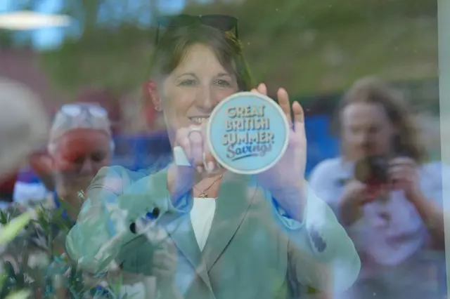 Reeves sticking a sticker which says 'great british summer savings' to the wall with people taking pictures behind her