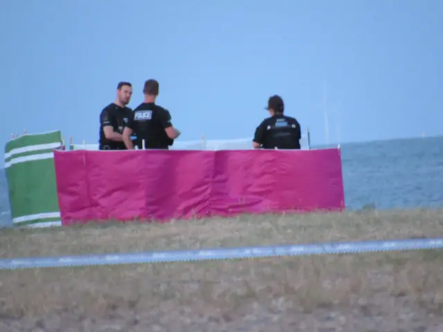 Police stand in between some wind breakers on the beach.
