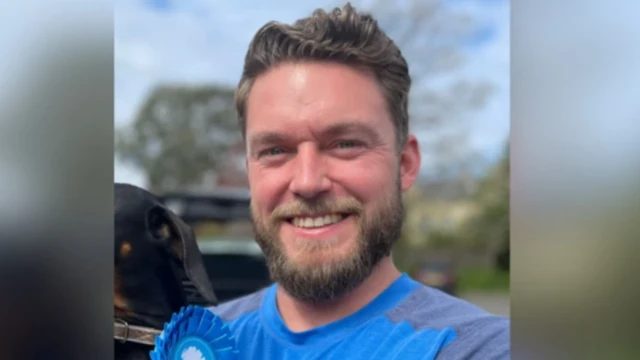 Phil Eckersley has dark hair and a beard. He wears a blue t-shirt and a matching Tory rosette