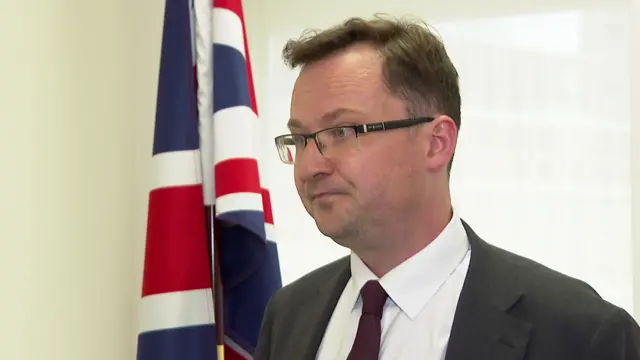 Justice Secretary Alex Norris in a dark grey suit, white shirt and burgundy tie listens to questions from a reporter (off camera) while standing next to a Union flag