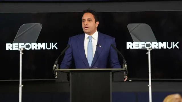 Reform UK's Zia Yusuf delivering a speech from behind a black lectern. Behind him, two screens broadcasting the party logo (Reform UK, in white, with a black arrow serving as the central hole in the letter O)