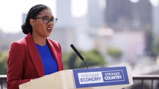 Kemi Badenoch wears a red blazer and a blue top as she delivers a speech behind a podium
