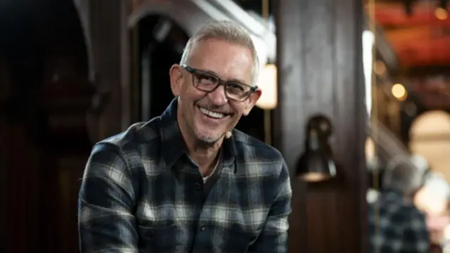 Gary Lineker wears a checkered shirt and glasses and smiles at the camera