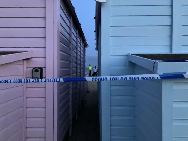 Police tape around a beach hut, with police officers in the distance