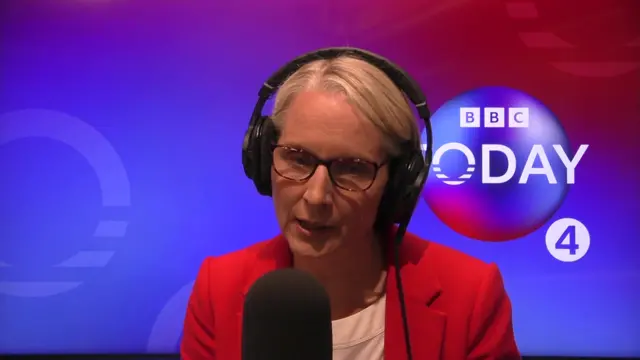 Emma Reynolds in a red blazer and white shirt speaking into a black mic. She has short blond hair held back by large radio headphones
