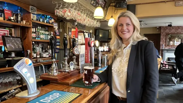 Emma McClarkin stands in a pub with a pint of beer. She is wearing a dark blazer and light blouse.