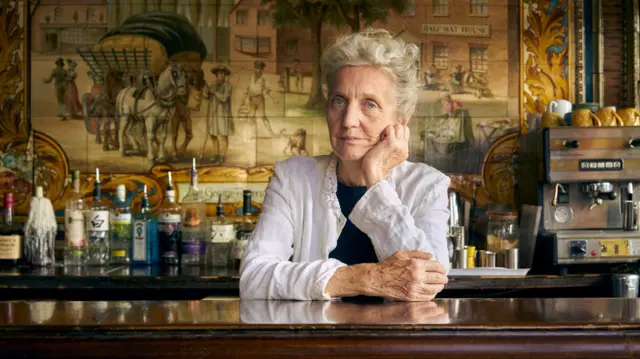 Pauline Forster stands behind the bar of the George Tavern, with tiles visible on the wall behind her.