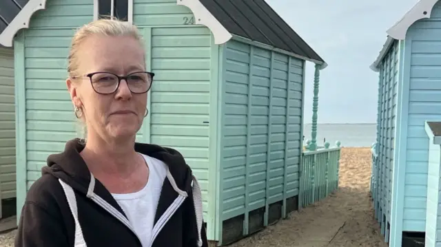 A woman with blonde hair and wearing glasses is standing in front of colourful beach huts. Behind her is some sand and a wooden railing. She is wearing a black hoody over a white top.